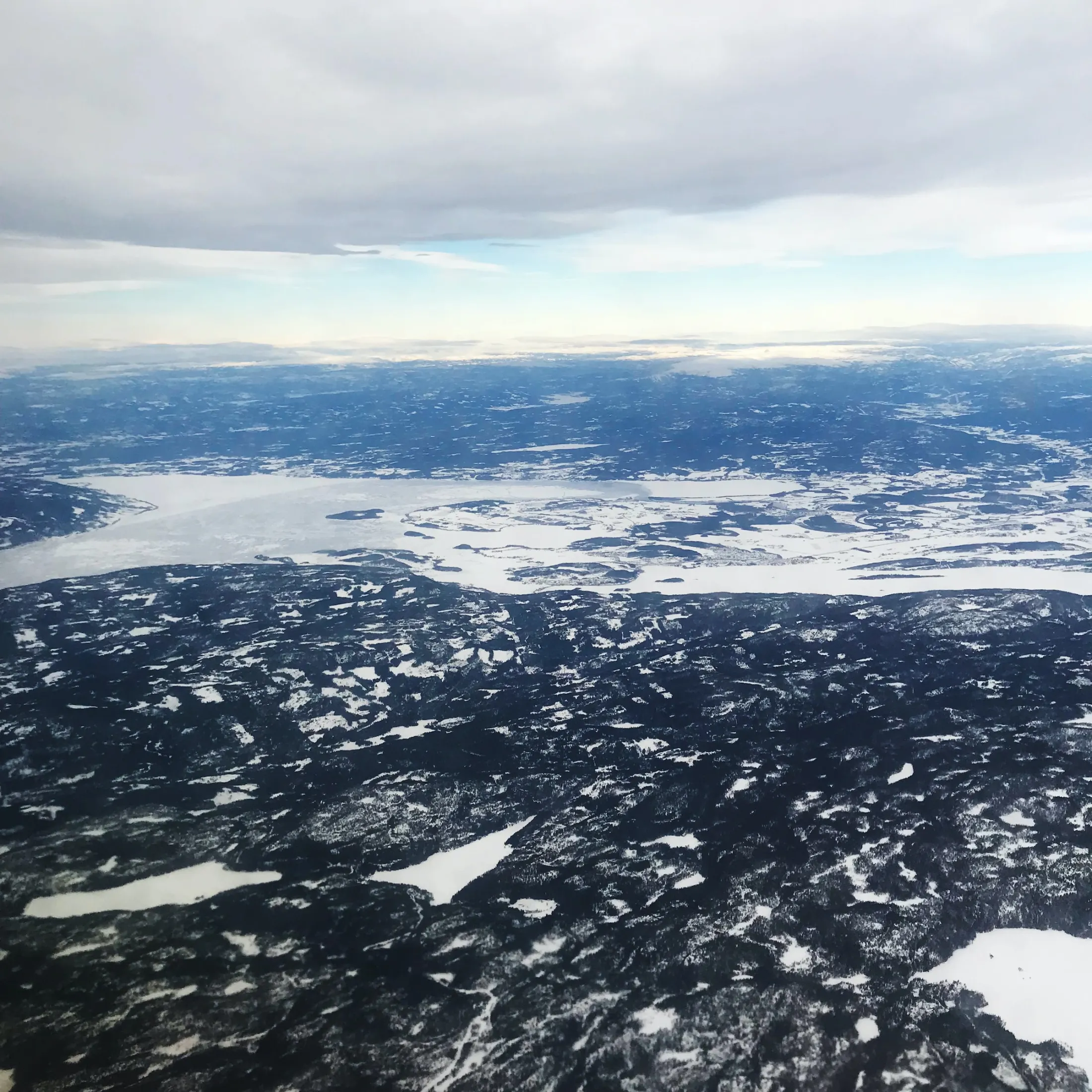 Luftfoto av et snødekt vinterlandskap med fjell og vann under en skyet himmel.