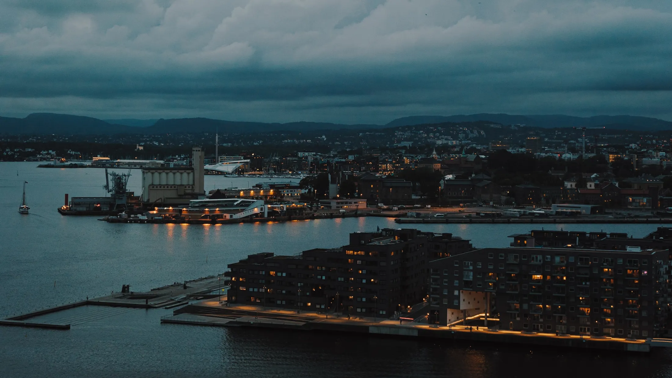 Oversikt over en kystby i skumringen, med lys som speiler seg i vannet og mørke skyer over.