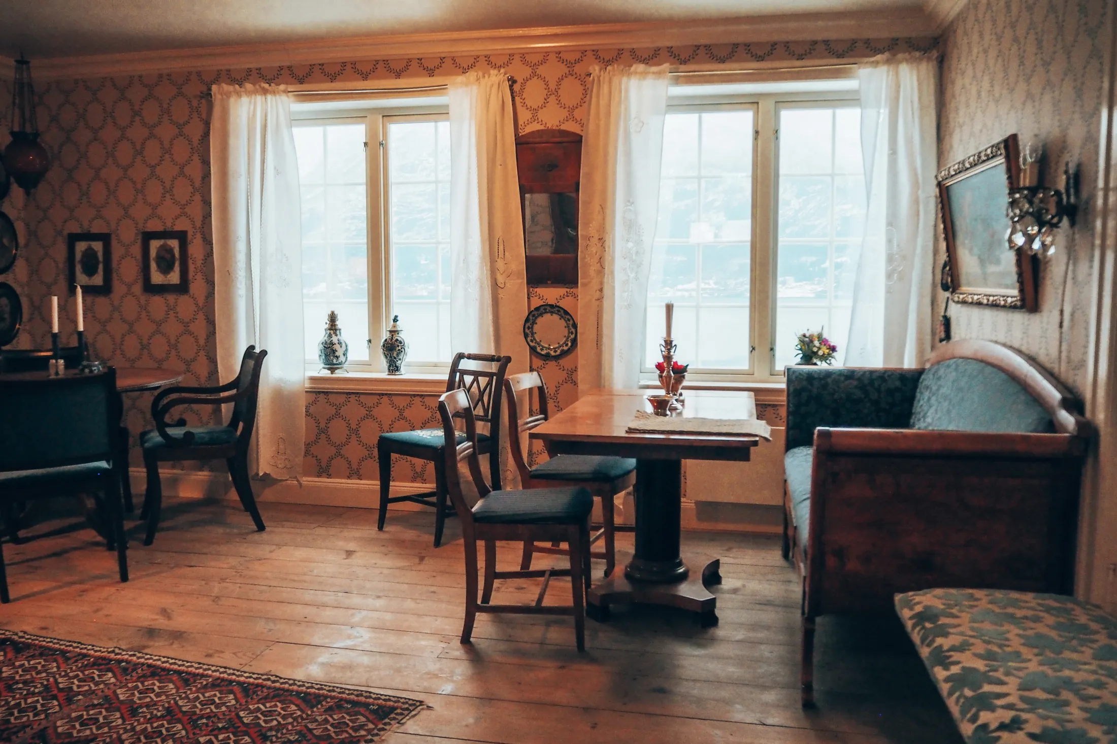 Et tradisjonelt spiserom med tregulv, mønstret tapet og eldre tremøbler foran to store vinduer med hvite gardiner.