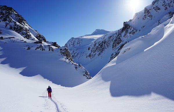 En ensom skiløper som går gjennom en snødekt fjelldal under en klar blå himmel.