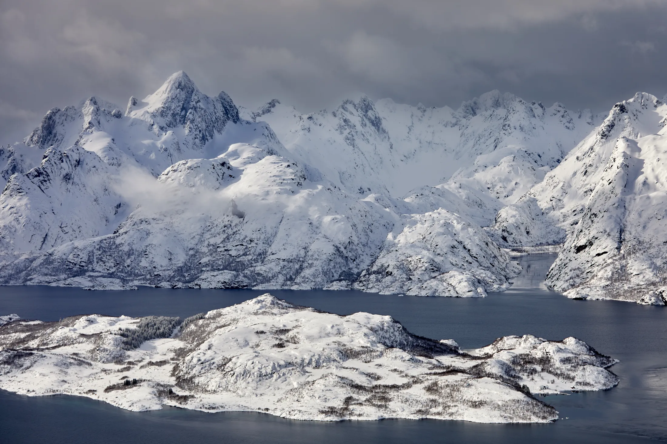 Snødekte fjell som reiser seg over en fjord med en snødekt øy i forgrunnen under en skyet himmel.