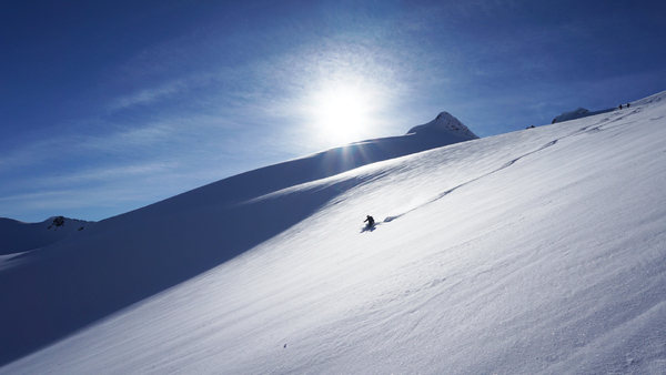 En ensom skiløper på en stor, snødekt fjellside under en sterk sol og blå himmel.