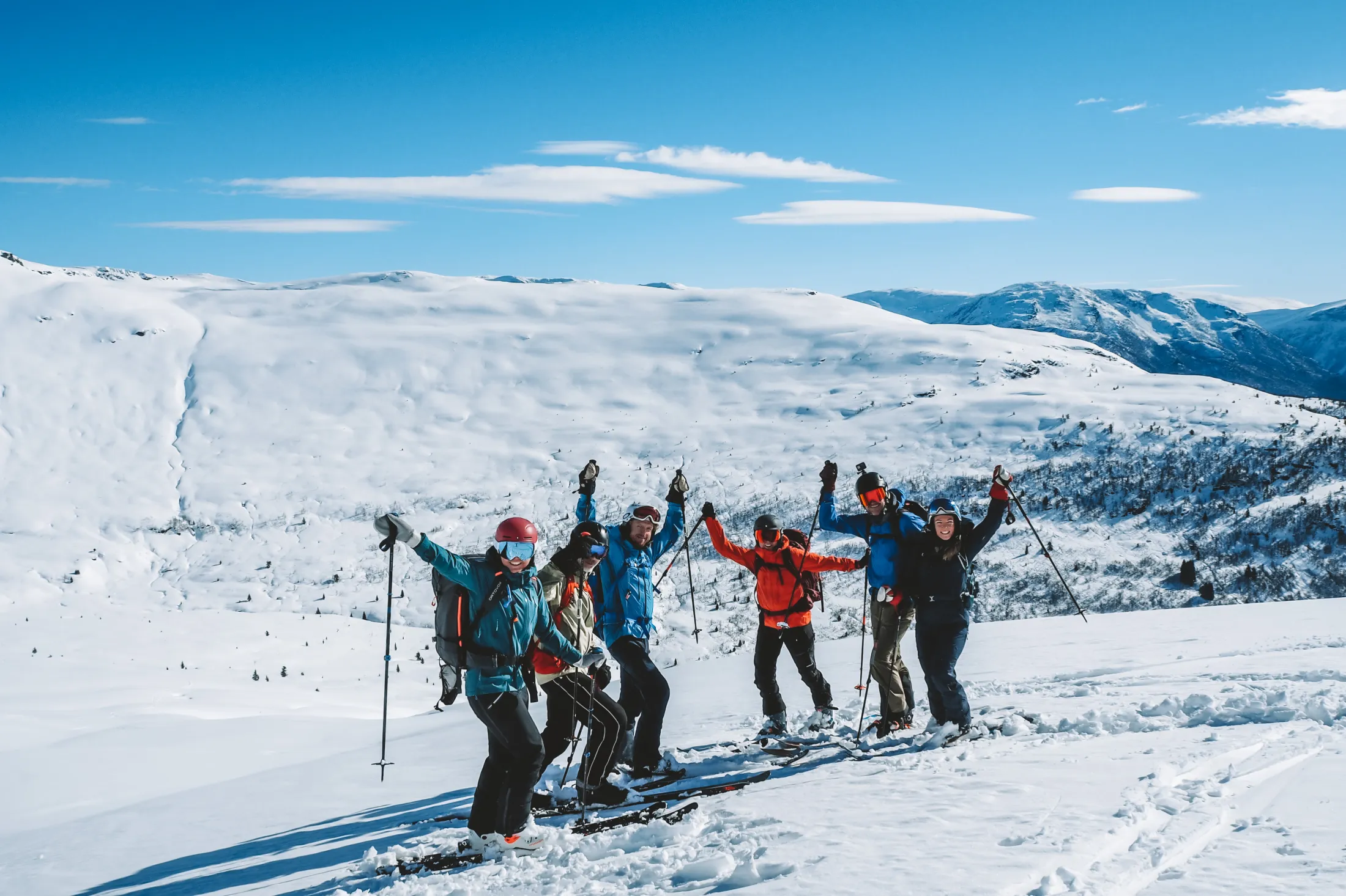 En gruppe glade skiløpere i fargerike vinterklær jubler med armene og stavene i været på en snødekt fjelltopp under en blå himmel.