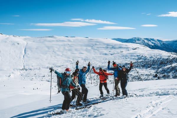 En gruppe glade skiløpere i fargerike vinterklær jubler med armene og stavene i været på en snødekt fjelltopp under en blå himmel.