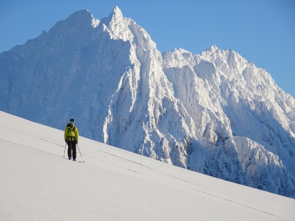 En ensom skiløper i skarp gul jakke går opp en stor snødekt skråning med ruvende snøfjell under en klar blå himmel i bakgrunnen.