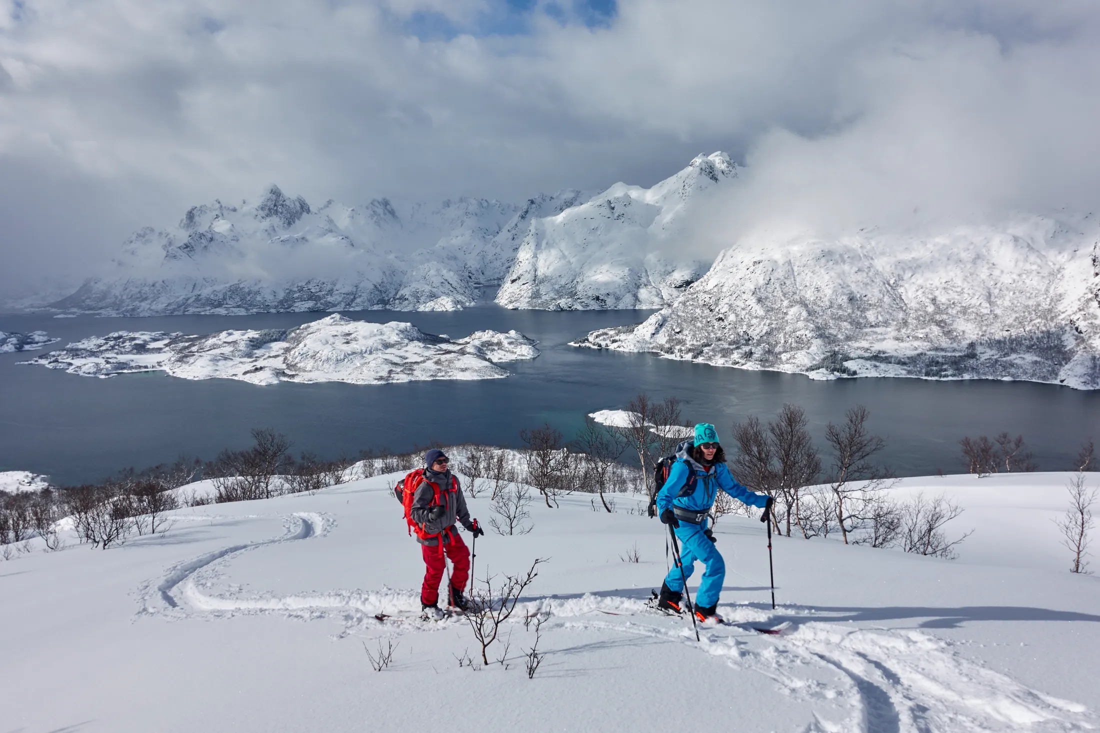 To skiturister som går oppover en snødekt skråning med utsikt over en fjord og fjell i Lofoten.