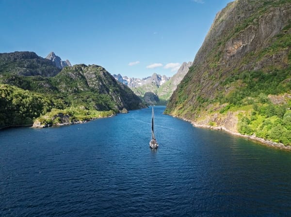 En seilbåt som navigerer i en smal, blå fjord omgitt av bratte, grønne fjell under en blå himmel.