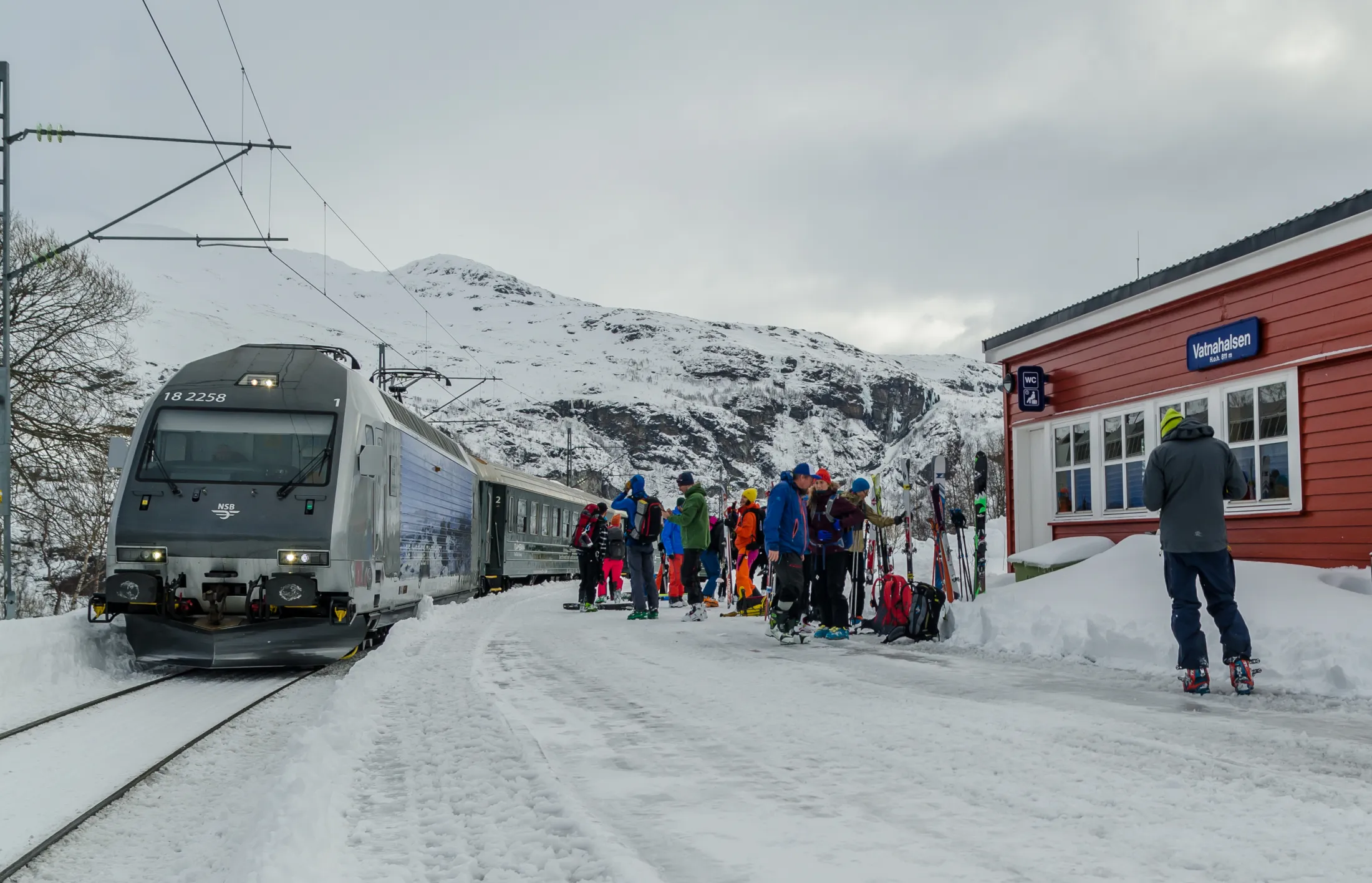 Et tog parkert på en snødekt stasjon med passasjerer på plattformen, og fjell i bakgrunnen.