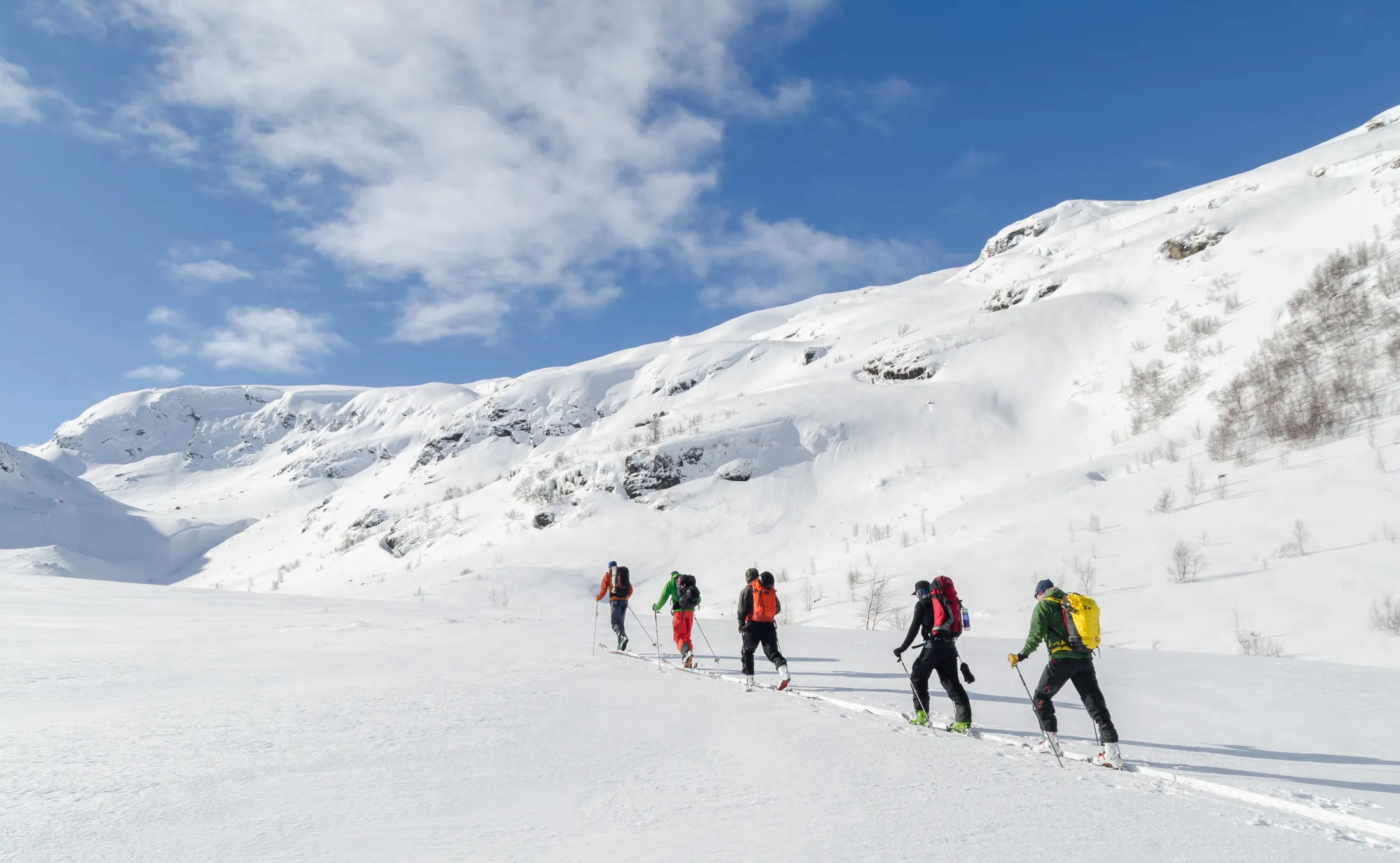 En rekke på fem skiløpere med sekk går på ski gjennom en snødekt dal mot bratte, snødekte fjell under en blå himmel med lette skyer.