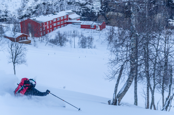 En skiløper med rød ryggsekk svinger seg gjennom dyp puddersnø i en helling med utsikt mot et rødt fjellhotell nede i dalen.