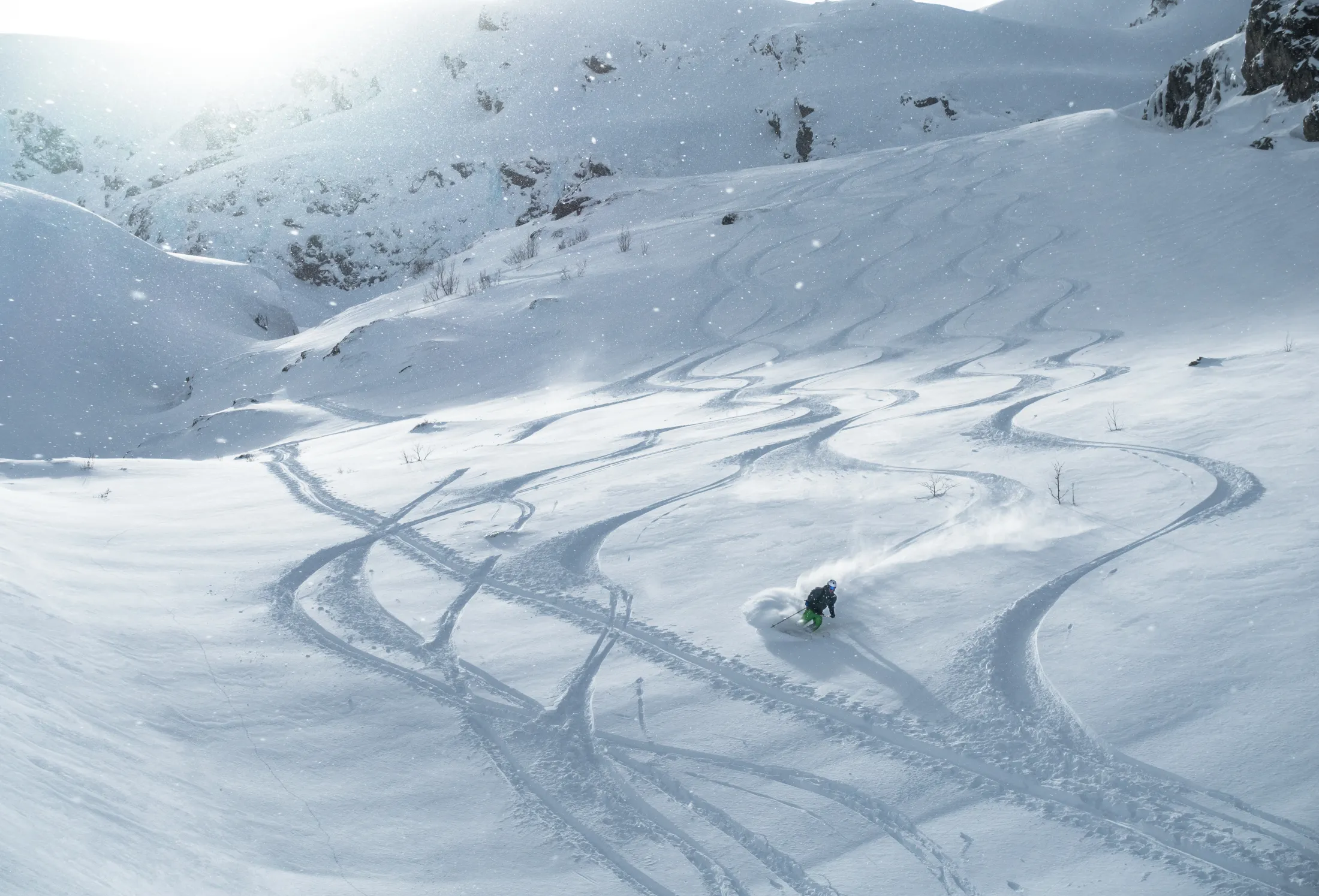 En enslig skiløper svinger seg gjennom dyp puddersnø i en bratt helling fylt med svingete skispor under sterk sol.
