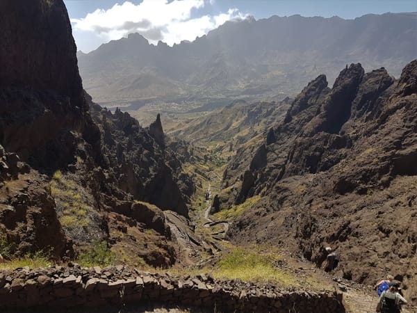 En turgåer går ned i en enorm, røff vulkansk canyon der en steinesti snor seg nedover mot en fjern dalbunn omkranset av ruvende fjelltopper.