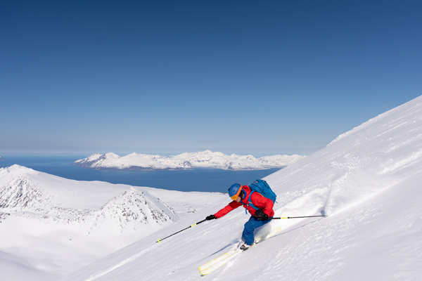 En skiløper i rød jakke kjører ned en snødekt skråning med utsikt over fjell og hav i bakgrunnen.