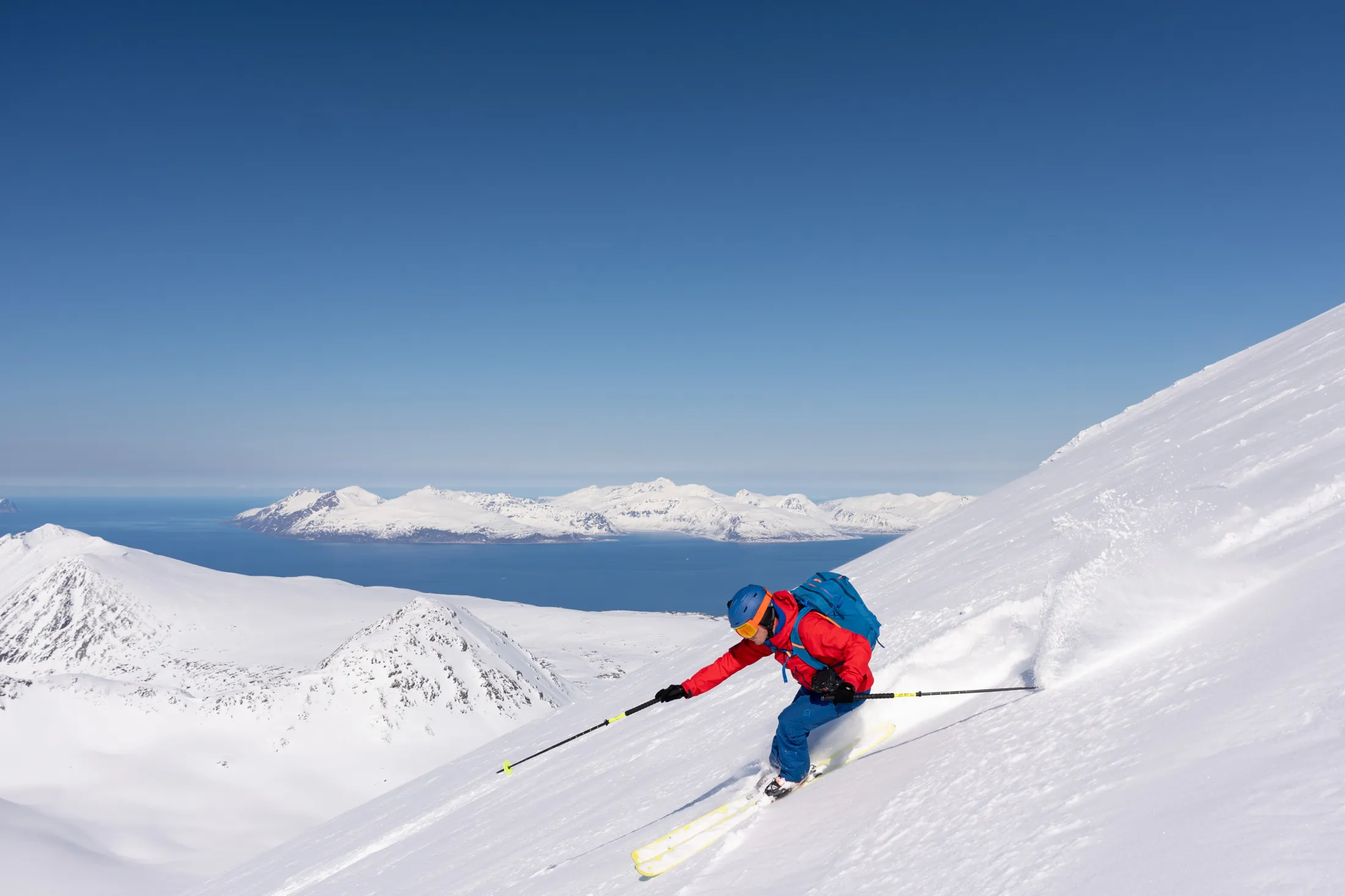 En skiløper i rød jakke kjører ned en snødekt skråning med utsikt over fjell og hav i bakgrunnen.