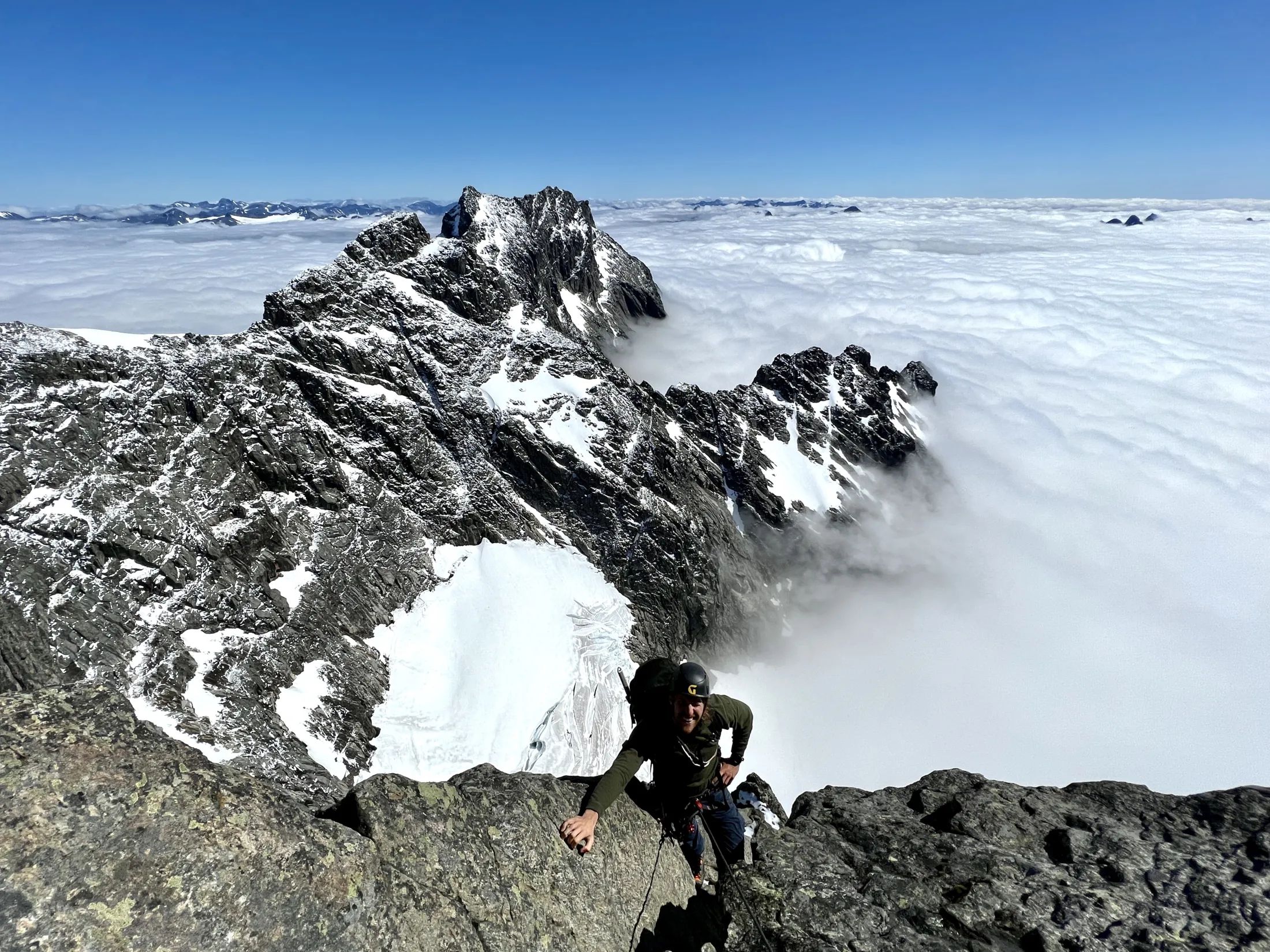 En klatrer som tar seg opp en forreven fjellkam over et tykt tåkehav under en klar blå himmel.