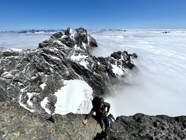 En klatrer som tar seg opp en forreven fjellkam over et tykt tåkehav under en klar blå himmel.