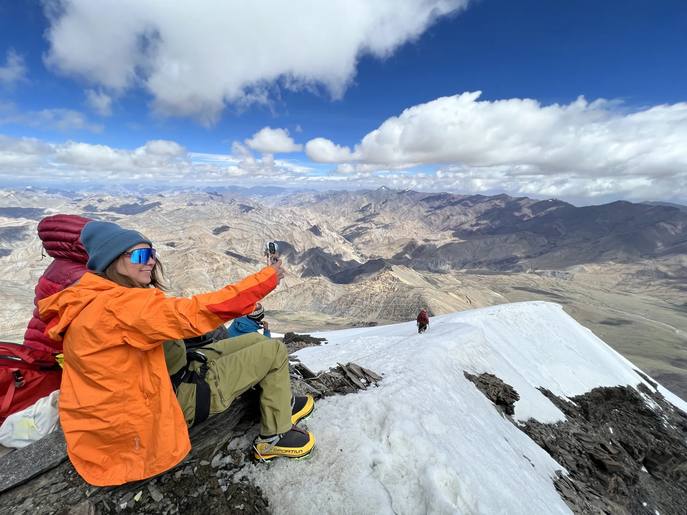 En fjellklatrer i en sterk oransje jakke sitter på en snødekt fjellrygg og tar en selfie, med et enormt, kupert fjellandskap som strekker seg ut under en delvis skyet blå himmel.