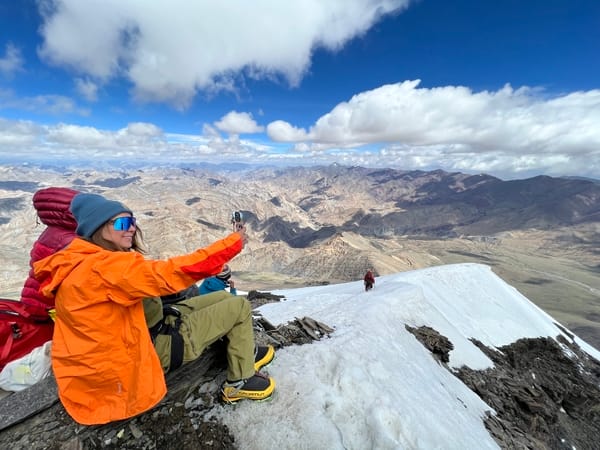 En fjellklatrer i en sterk oransje jakke sitter på en snødekt fjellrygg og tar en selfie, med et enormt, kupert fjellandskap som strekker seg ut under en delvis skyet blå himmel.