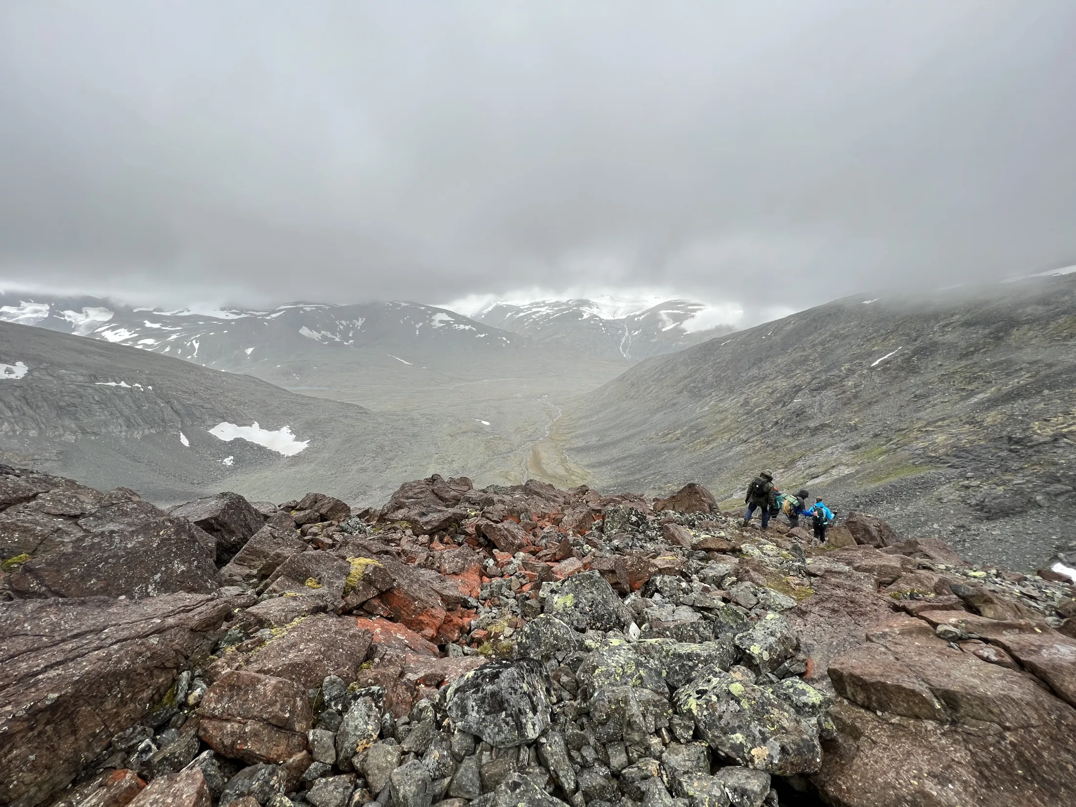 Fjellvandrere går langs en steinete fjellrygg med utsikt over en tåkete dal under en overskyet himmel.