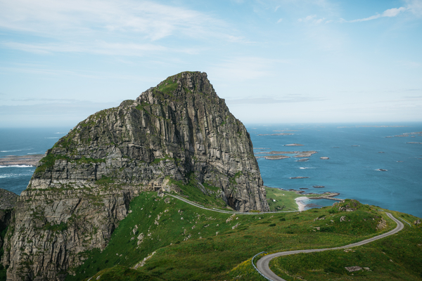 Et bratt, steinete fjell som reiser seg over grønne skråninger og det blå havet, med en svingete vei i forgrunnen.