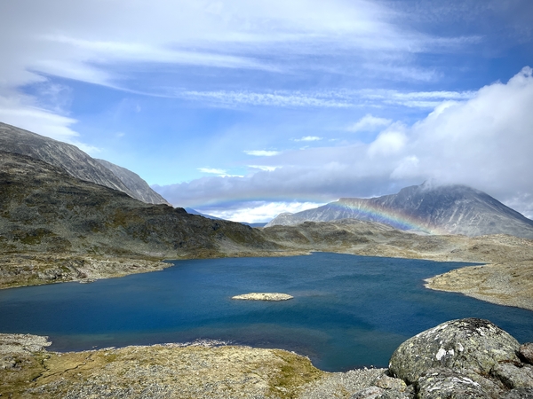 Et dypt blått fjellvann omgitt av et kupert og steinete landskap under en delvis skyet blå himmel.