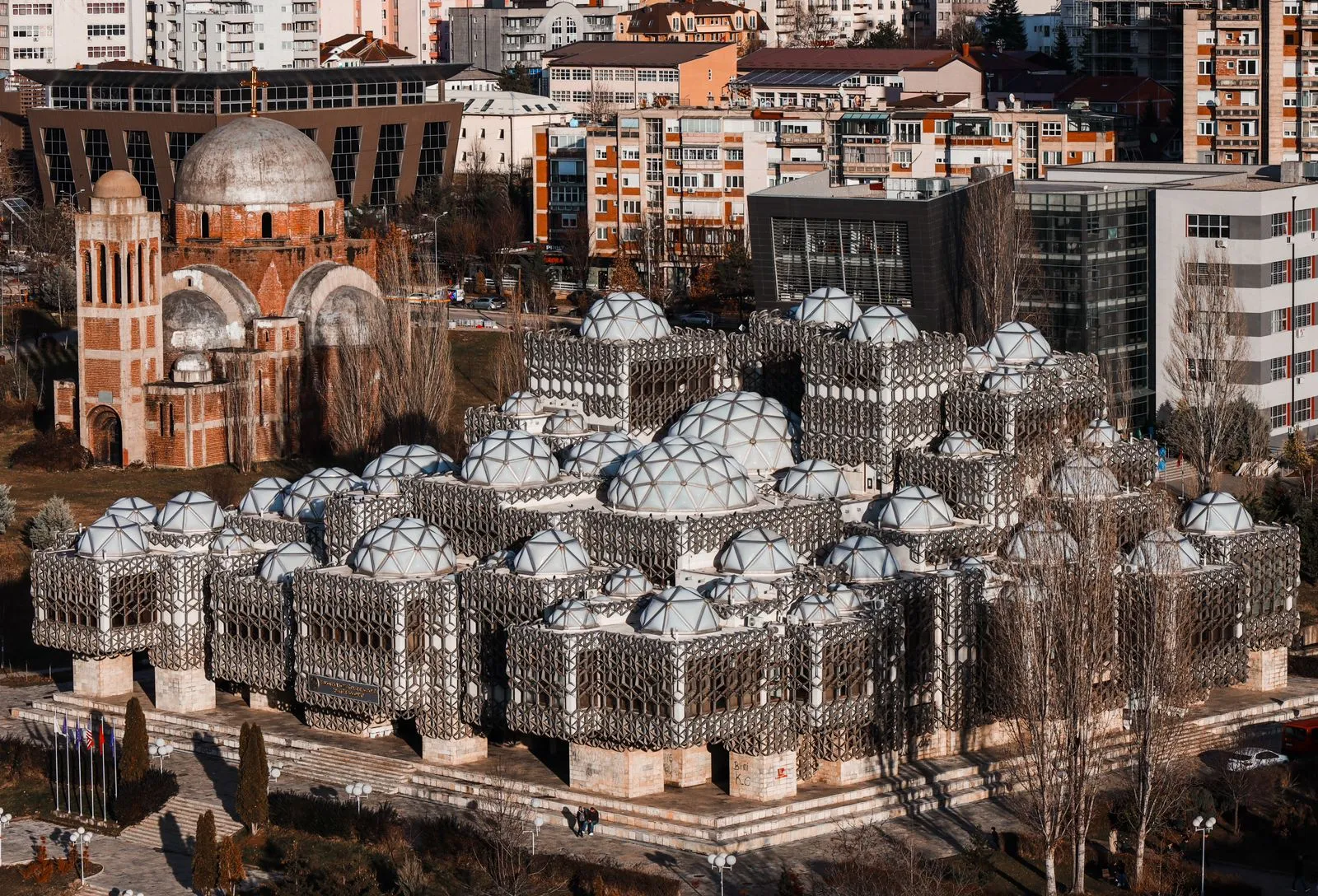 Et flyfoto av Kosovos nasjonalbibliotek i Pristina, som viser den unike brutalistiske arkitekturen med en rekke små kupler og en geometrisk metallgitterfasade.