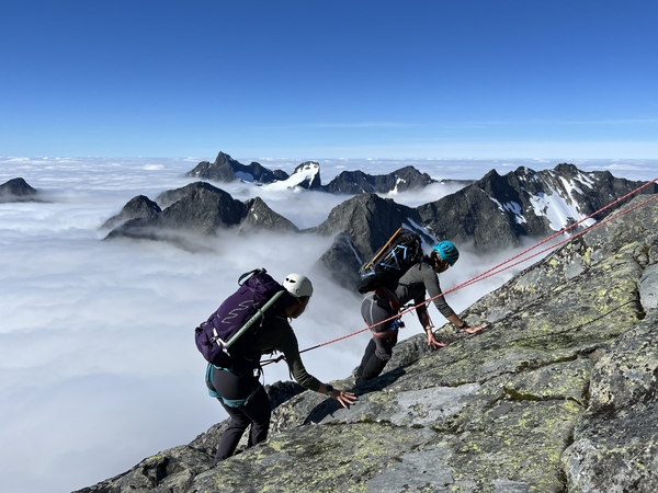 To fjellklifrere som klatrer på en steinete eg over et skyhav.