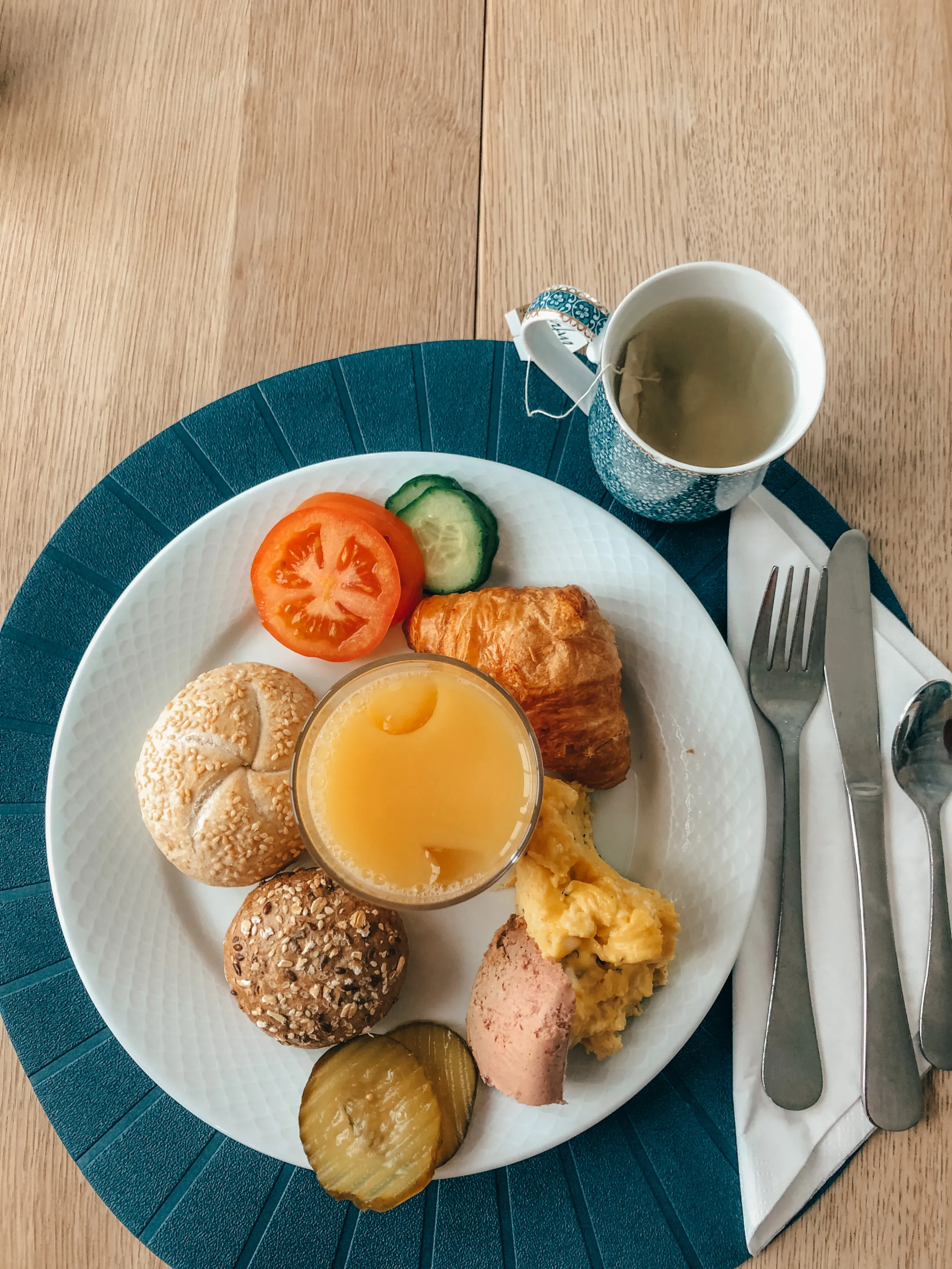 En frokosttallerken med rundstykker, croissant, eggerøre, tomat, agurk, sylteagurk og et glass juice, servert ved siden av en kopp te.