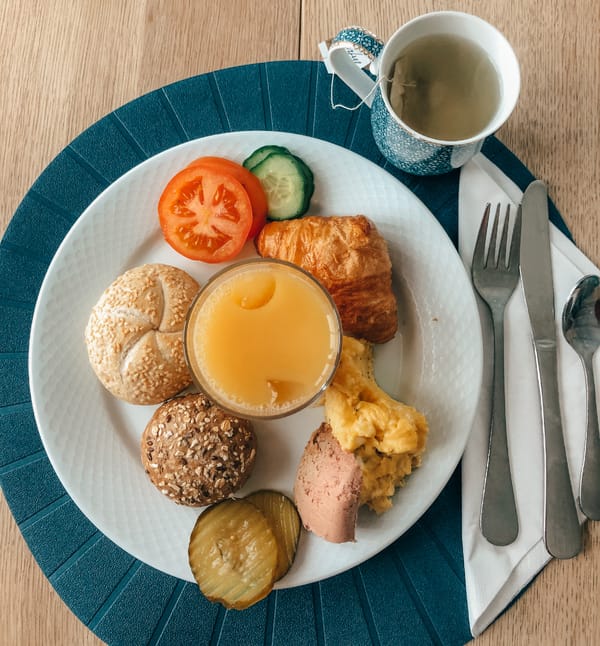 En frokosttallerken med rundstykker, croissant, eggerøre, tomat, agurk, sylteagurk og et glass juice, servert ved siden av en kopp te.
