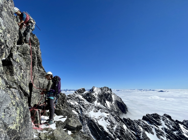 To klatrere på en bratt, snødekt fjellrygg under en klar blå himmel, med et hav av skyer under seg.