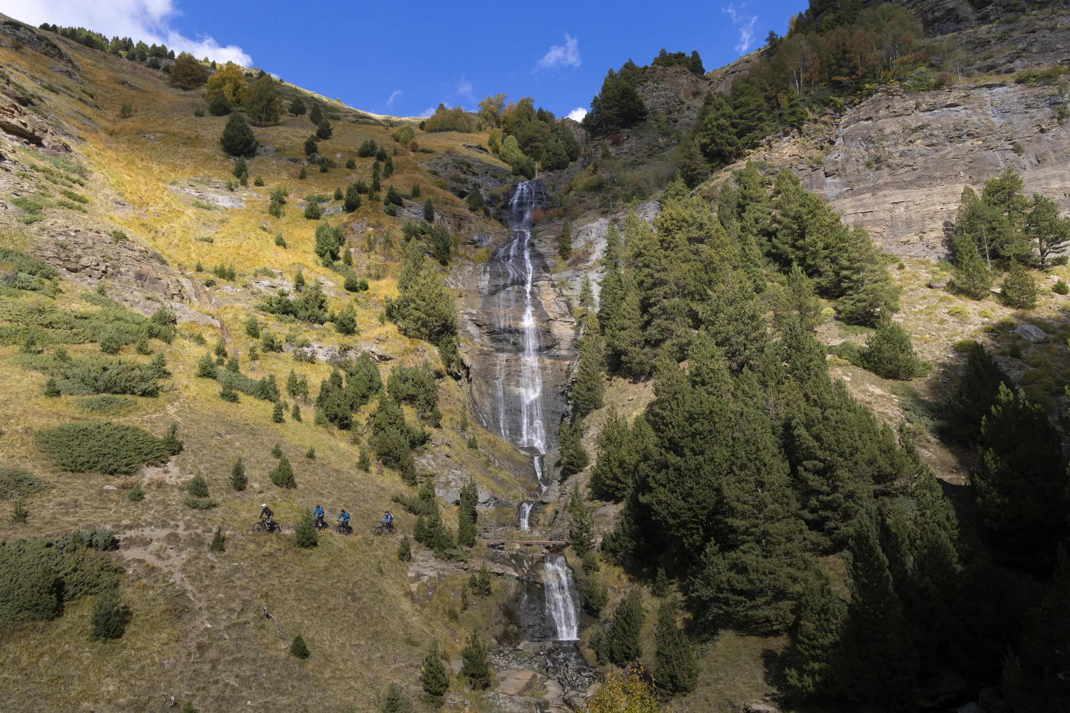 En høy foss som renner nedover en steinete fjellskråning med spredte trær.