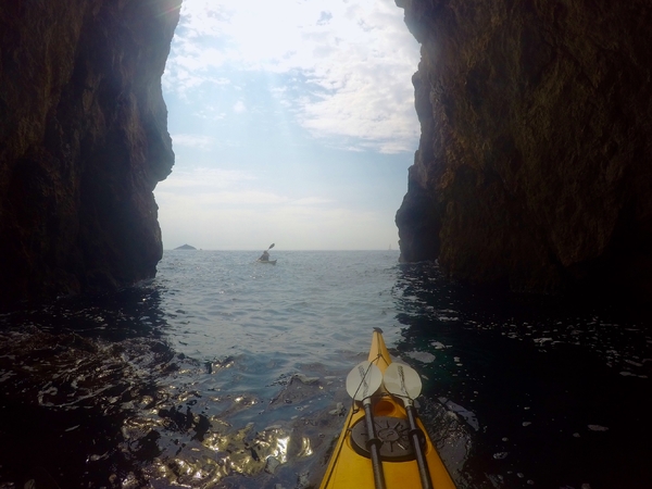En førstepersonsvisning fra en gul kajakk inne i en mørk sjøgrotte, med utsikt ut gjennom den høye fjellåpningen mot en annen kajakkpadler på det åpne havet under en lys, lettskyet himmel.