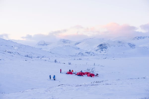 En ekspedisjonsleir med røde telt slått opp i et vidstrakt, snødekket fjellandskap under en lys morgenhimmel.