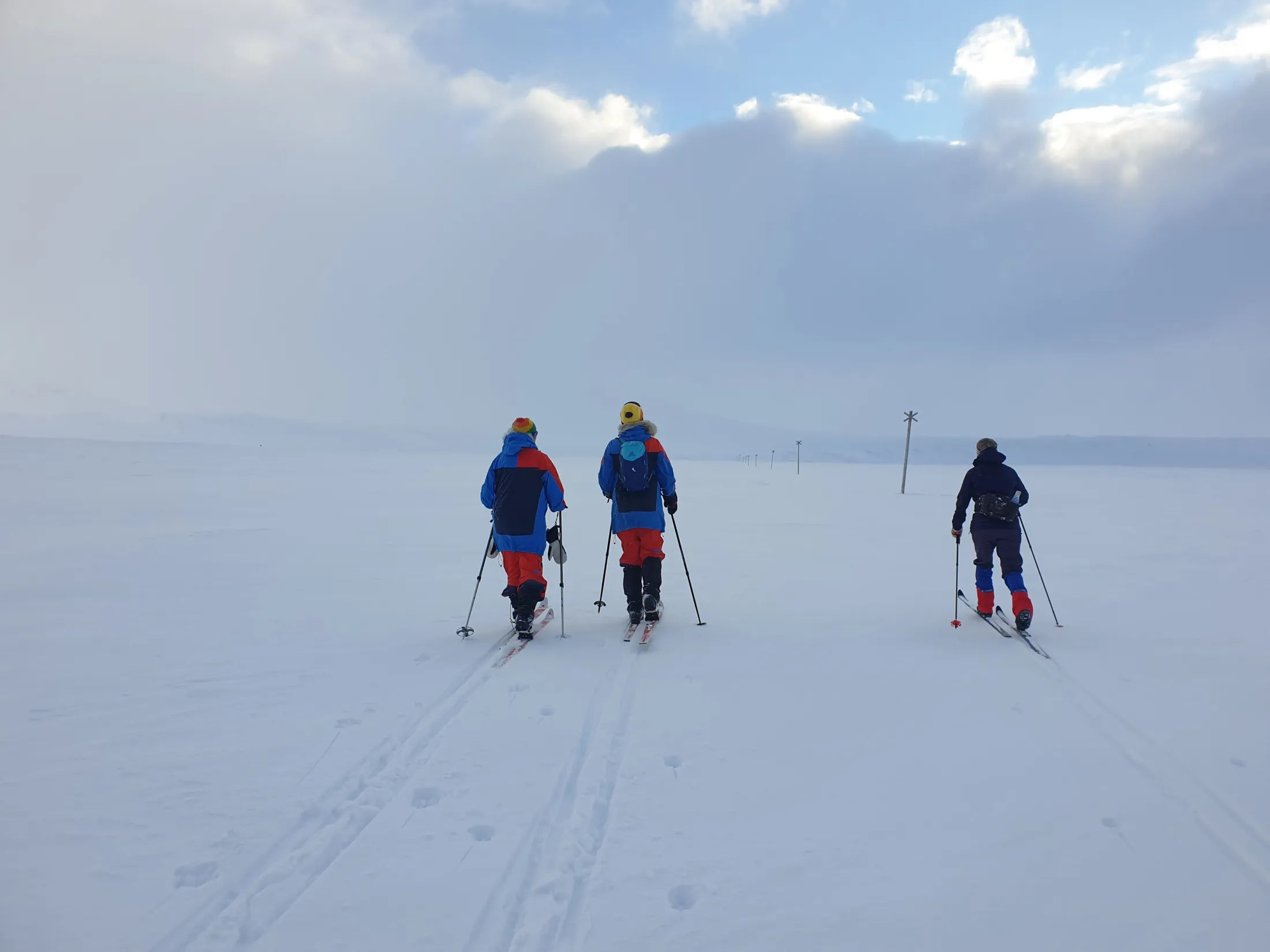 Tre langrennsløpere som går på ski over et stort, snødekt landskap under en skyet himmel.