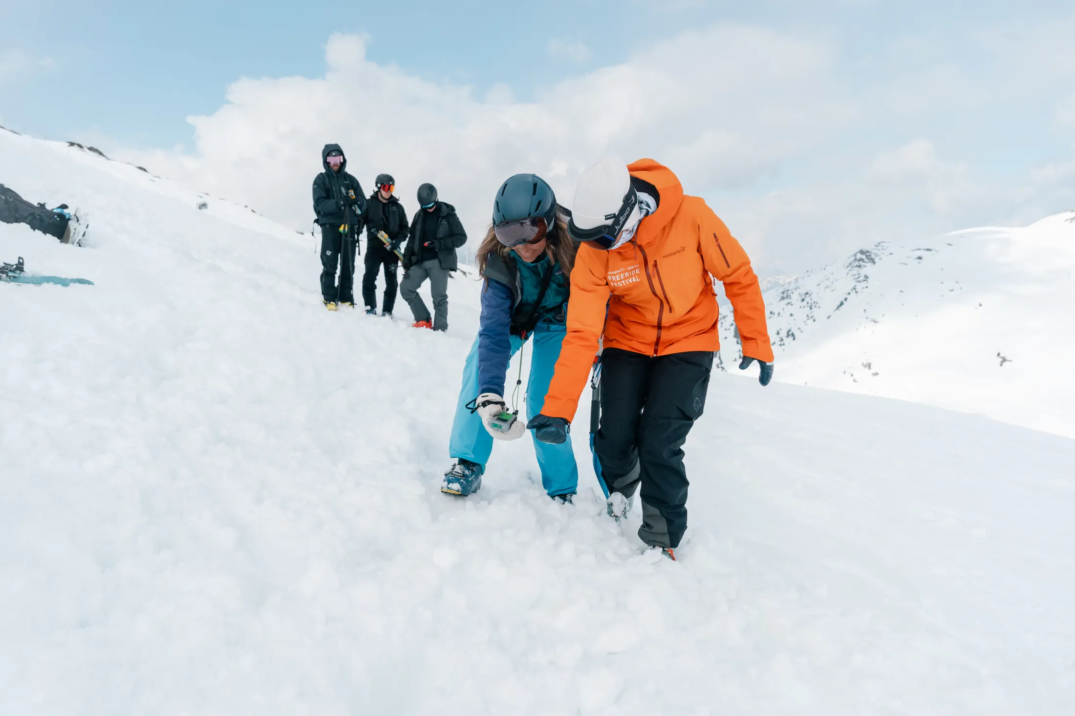 To personer i snøen, en blå og en orange jakke, går med skredsøker foran seg og øver på å bruke den. Tre mennesker står bak til venstre i skitøy. 
