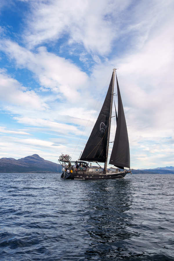 En seilbåt med svarte seil seiler på en fjord under en skyet blå himmel, med fjell i det fjerne.