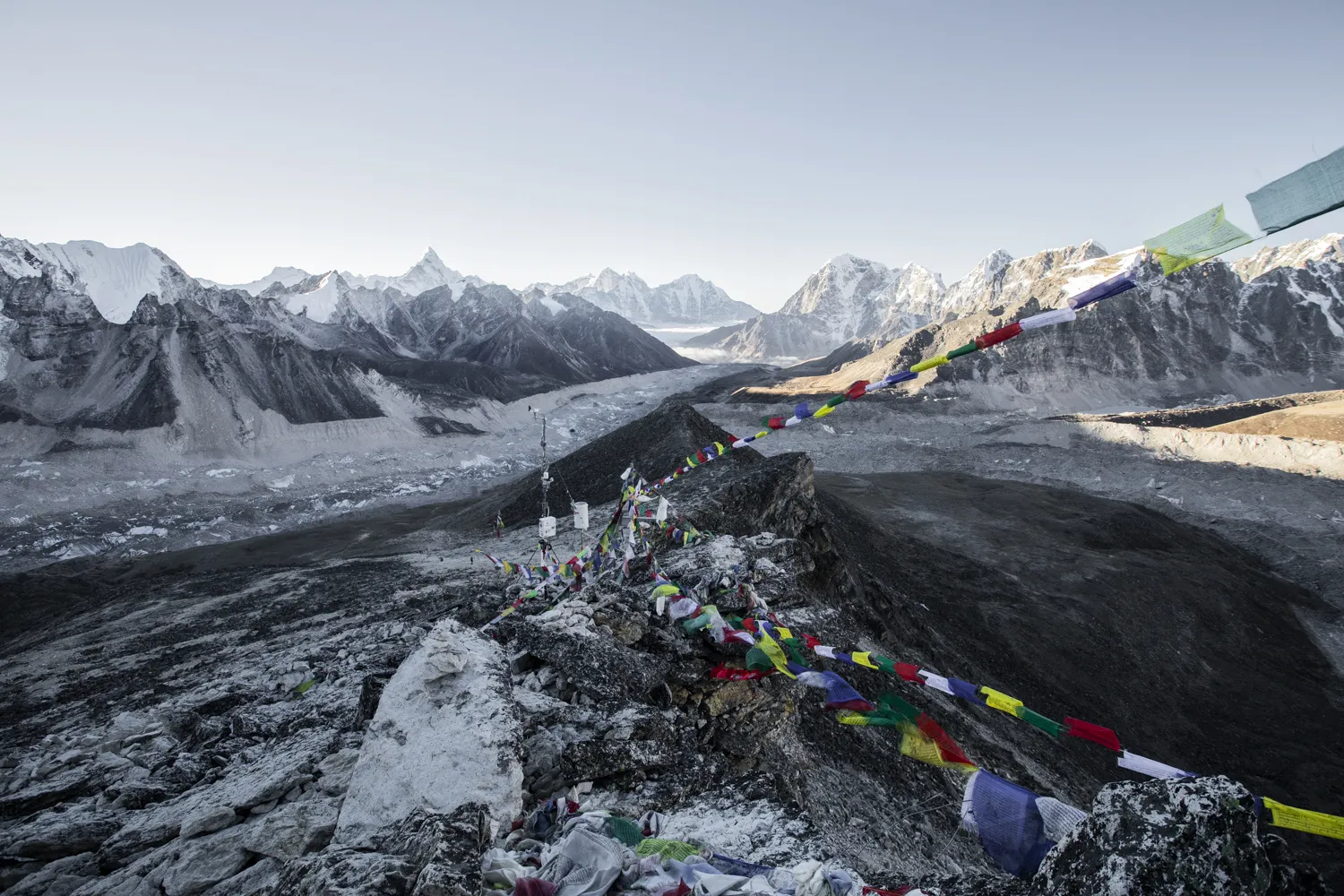 Høyt oppe på en steinete, snødekket rygg blafrer fargerike tibetanske bønneflagg i vinden, med utsikt over en enorm bredal som strekker seg mot en panoramisk horisont av spisse, snødekte himalayatopper.
