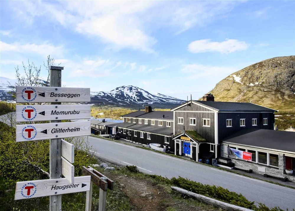 Et treskilt som viser vei til ulike stier i forgrunnen, med en fjellstue og snødekte topper i bakgrunnen.