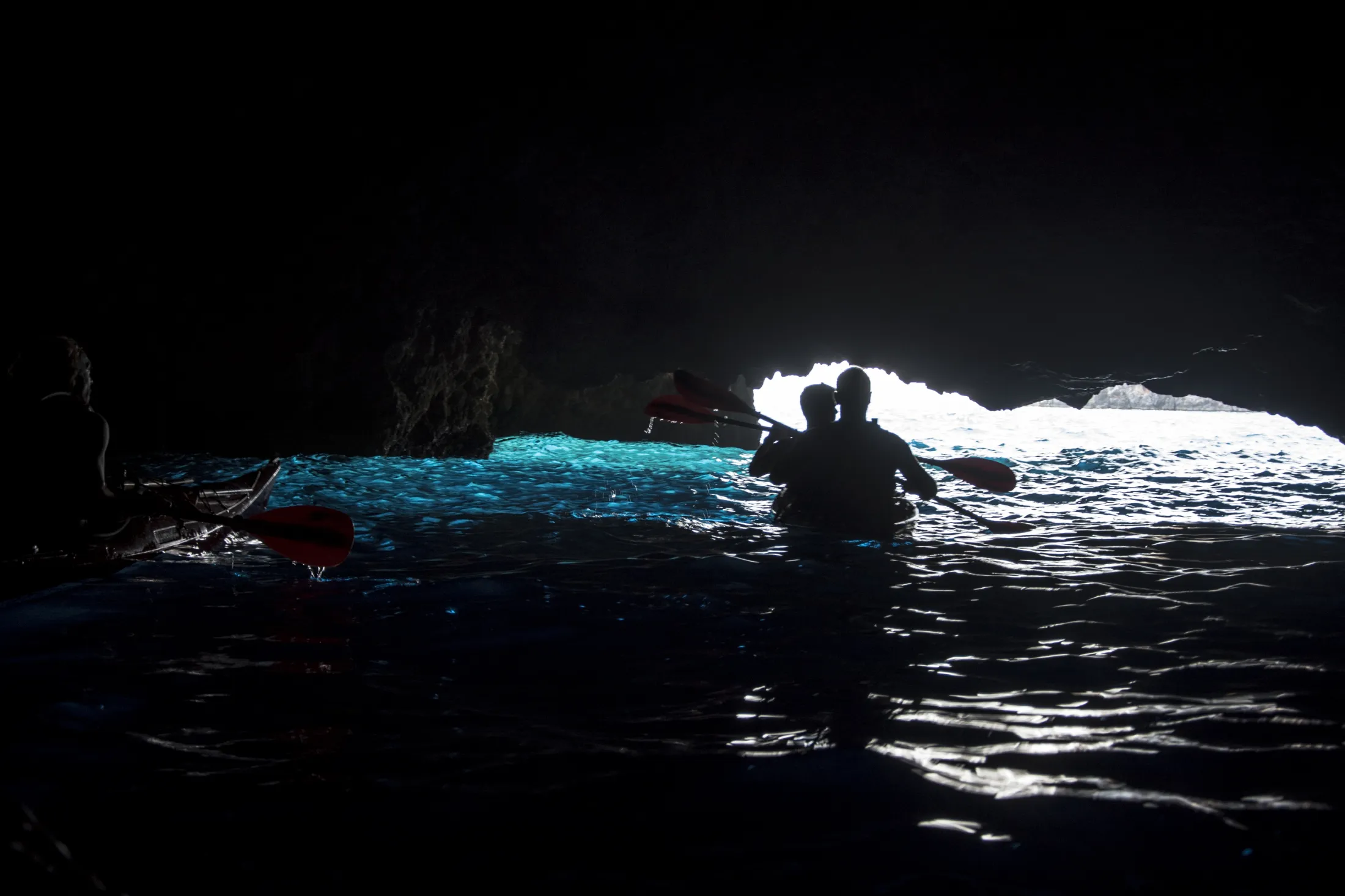 Silhuettene av kajakkpadlere med røde årer vises inne i en mørk sjøgrotte, mens de ser ut mot en lys utgang der det glødende turkisblå vannet reflekteres under fjelltaket.