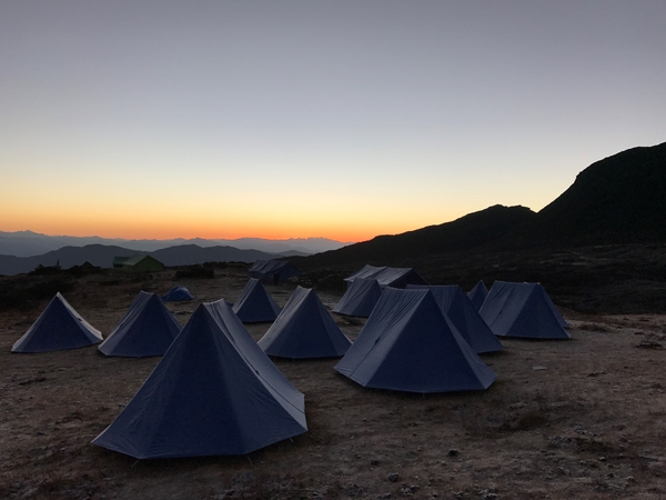 En samling blå spisstelt på en teltplass høyt til fjells under en fargesterk, oransje solnedgang.