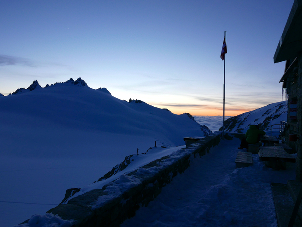 Et snødekt fjellandskap i daggry viser en steinterrasse med et sveitsisk flagg på en stang mot en fargerik horisont.