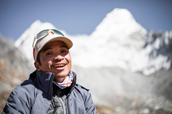 Et nærbilde av en smilende mann med caps og solbriller, med en vakkert uskarpt bakgrunn av snødekte fjelltopper mot en blå himmel.