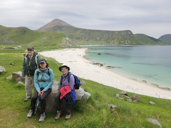 Tre turgåere poserer på en gresskledd åsside med en hvit sandstrand og turkis sjø i bakgrunnen.