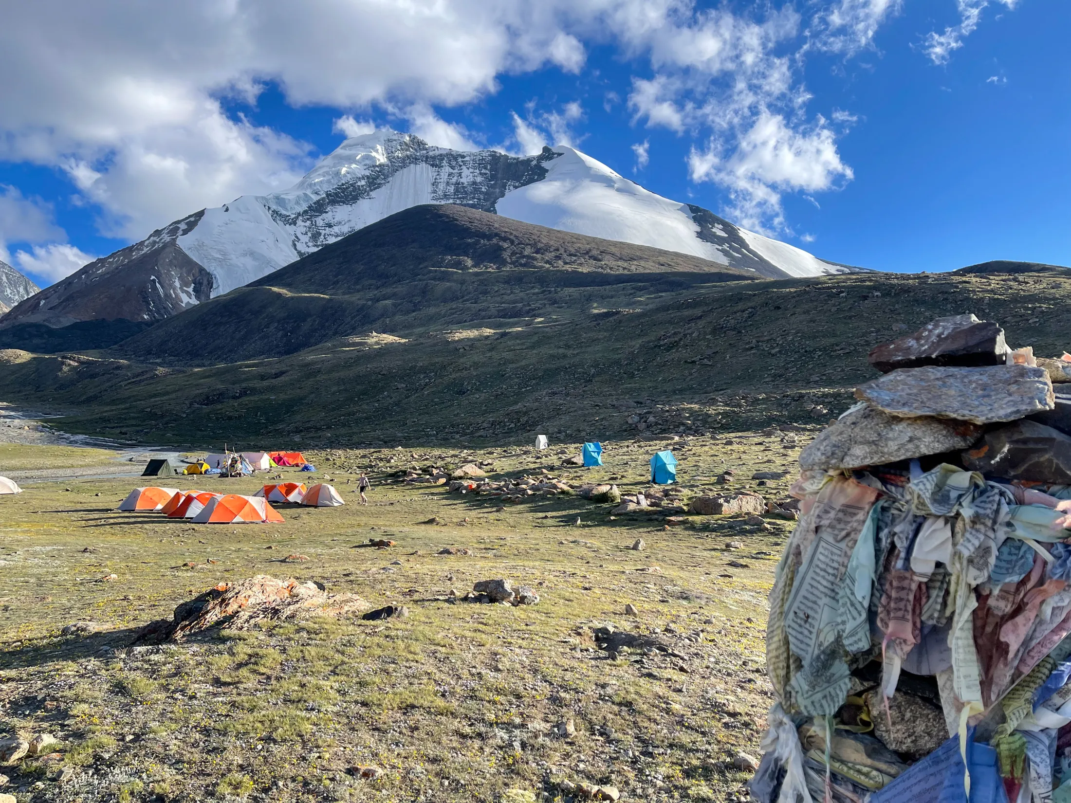 Flere oransje og blå telt er slått opp på en gresskledd teltplass ved foten av en massiv, snødekt fjelltopp under en delvis skyet blå himmel, med en steinvarde pyntet med bønneflagg i forgrunnen.