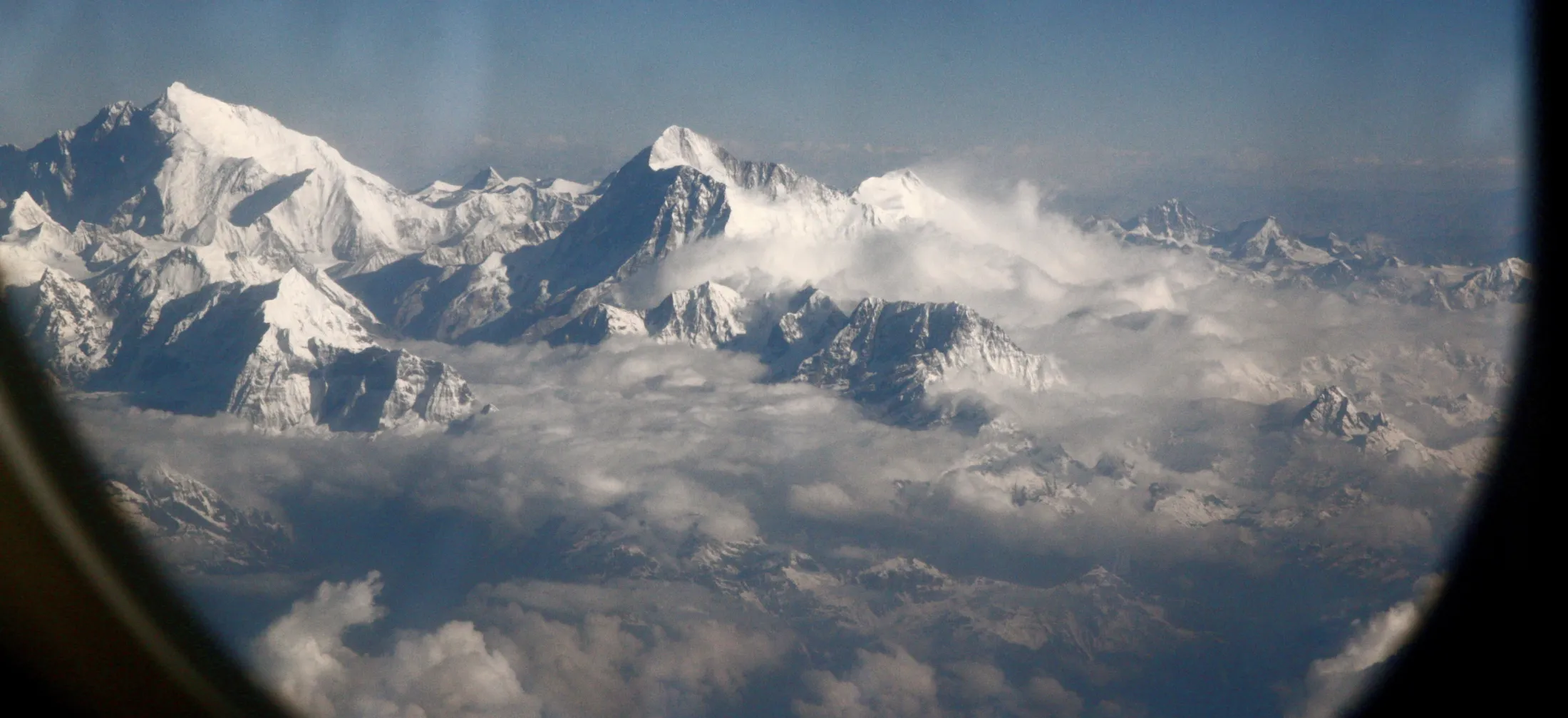 Utsikt fra et flyvindu som viser snødekte fjelltopper i Himalaya som stikker opp over et tykt skylag under en klar, blå himmel.