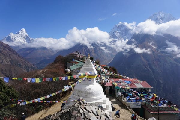 En hvit buddhistisk stupa dekorert med Buddhas øyne står i en fjellandsby, omgitt av fargerike bønneflagg og ruvende, snødekte fjell under en blå himmel.