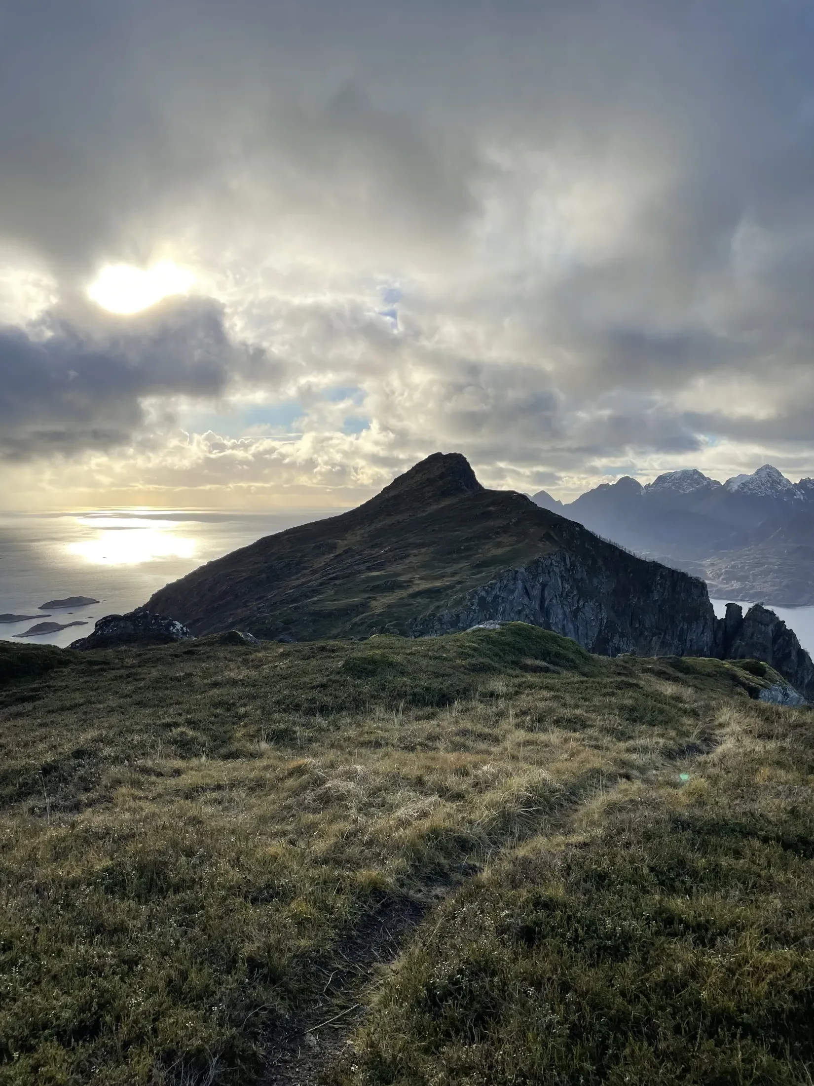 En dramatisk fjelltopp under en skyet himmel med sollys som bryter gjennom, fotografert fra en gressbakke.
