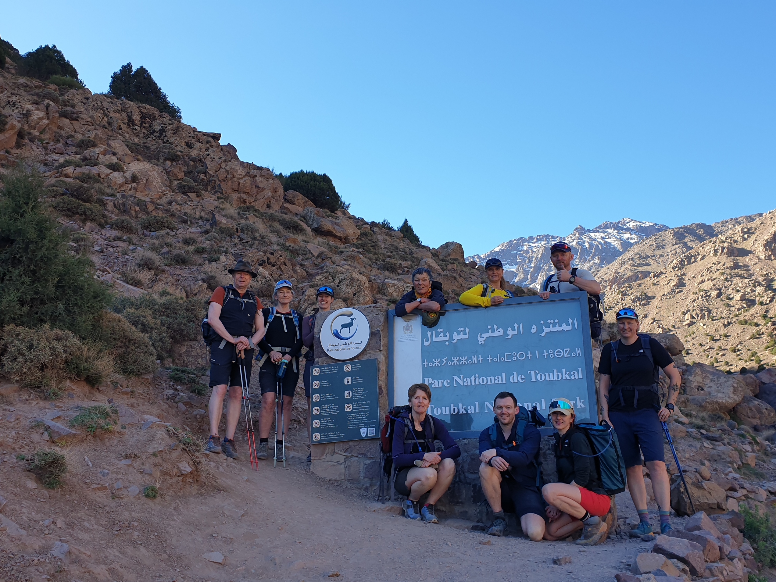 Folk som skal på tur står ved et skillt i fjellet med blå himmel