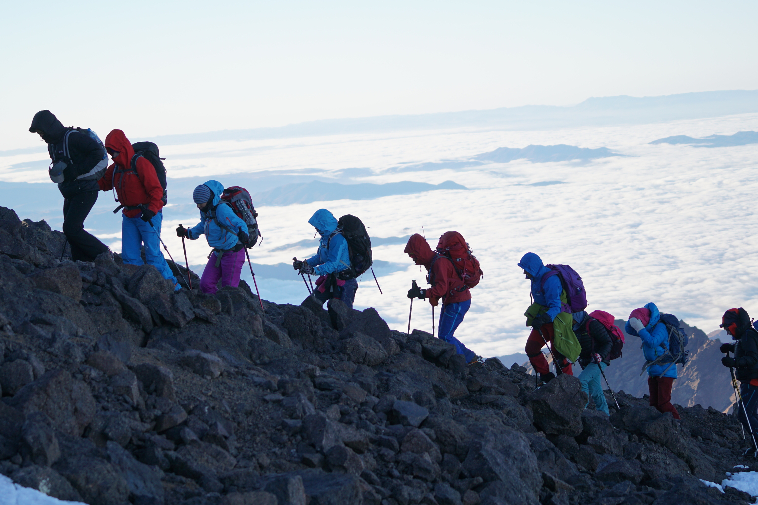 Folk som vandrer opp et fjell med mange klær i skyggen og skyer under seg i dalen